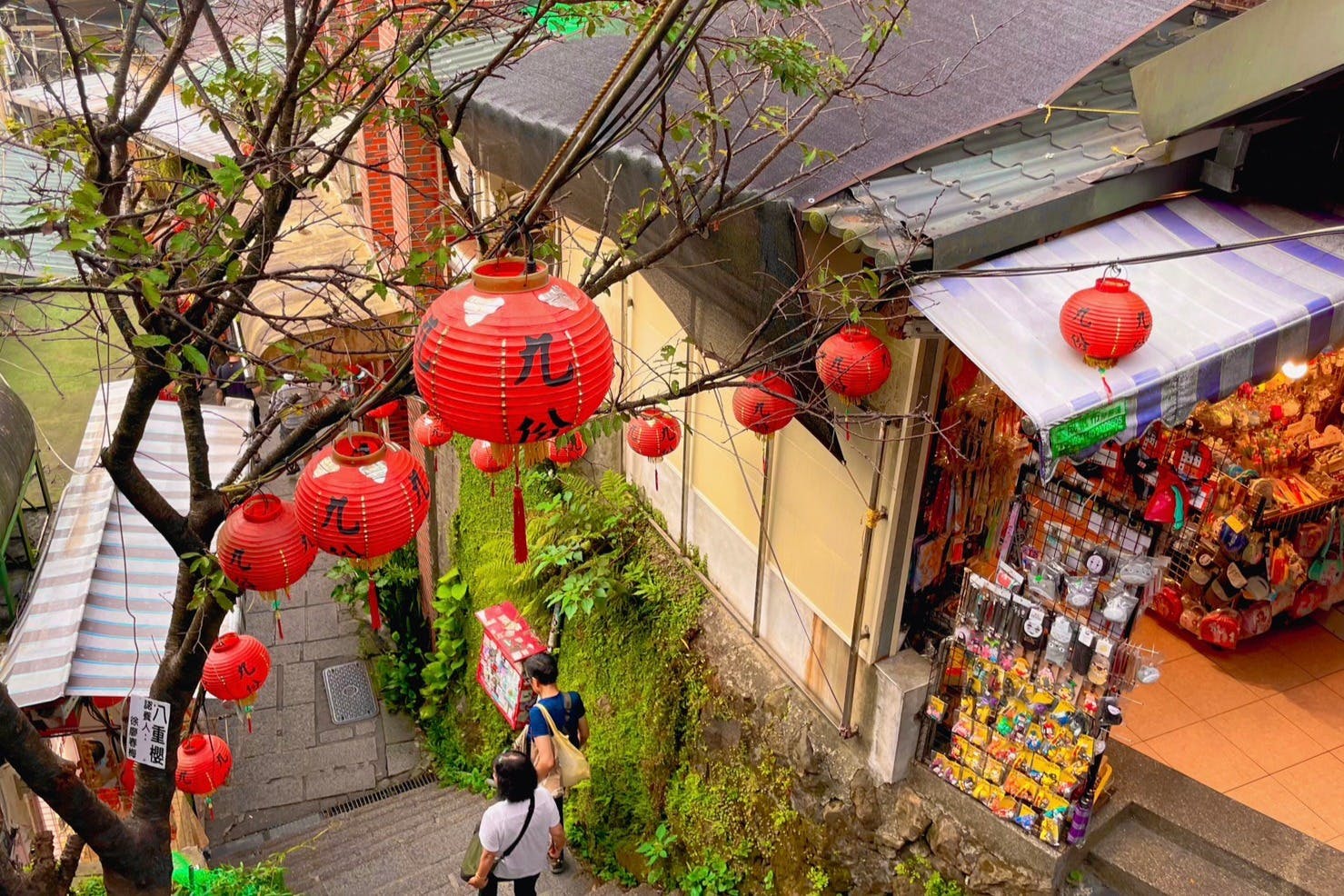 People walk down a narrow street adorned with red lanterns. A shop on the right displays various colorful trinkets.