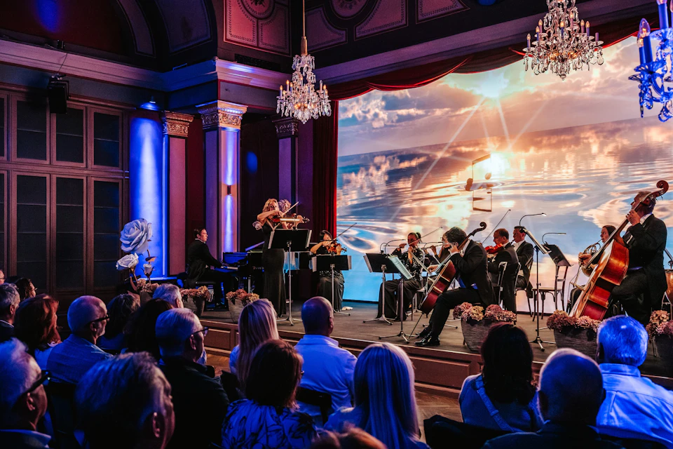 A string ensemble performing on a lit stage with an ocean-themed backdrop, viewed by an audience in a decorated hall.