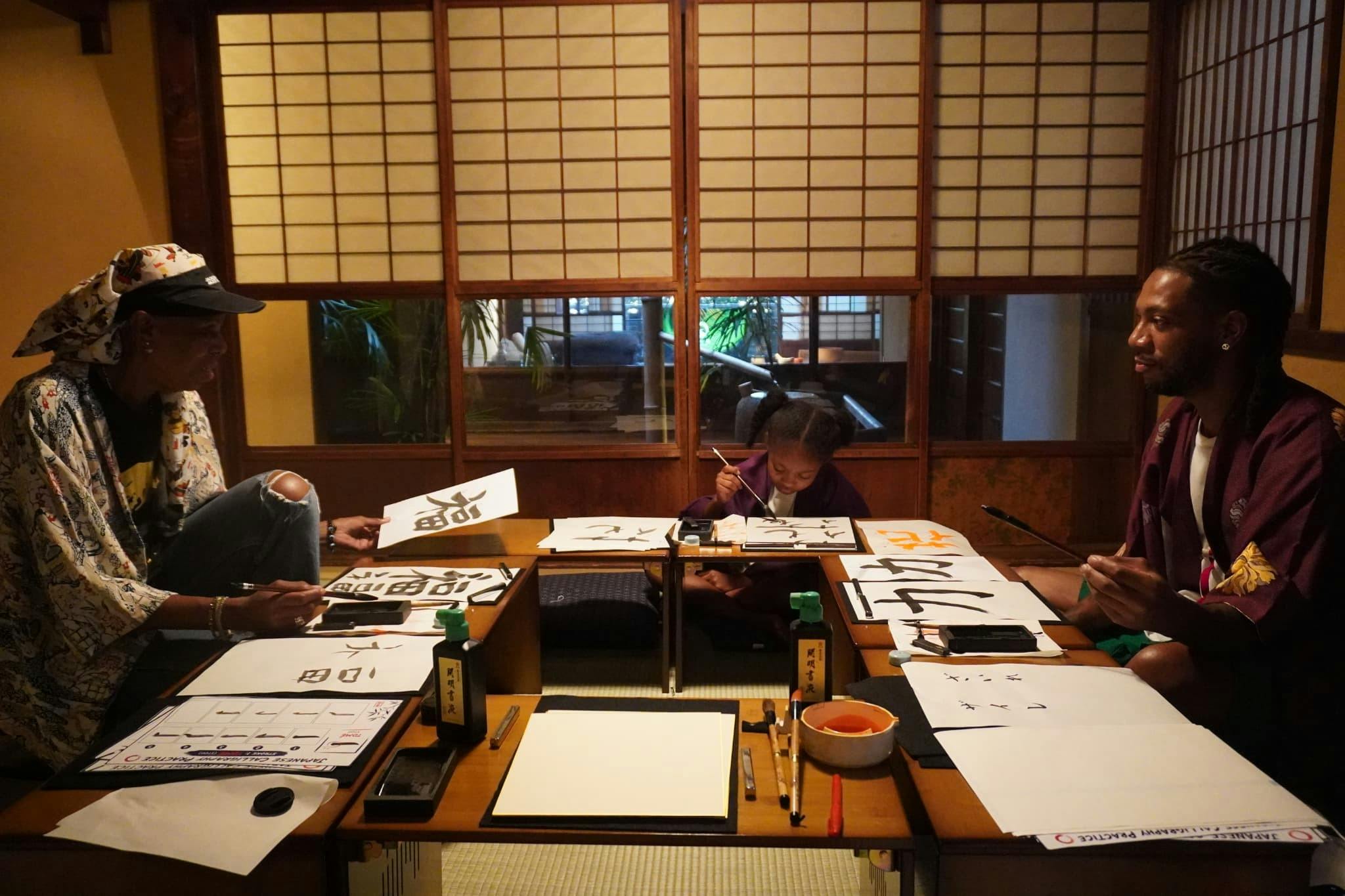 Two individuals practicing calligraphy in a traditional Japanese room with shoji screens and wooden furniture.