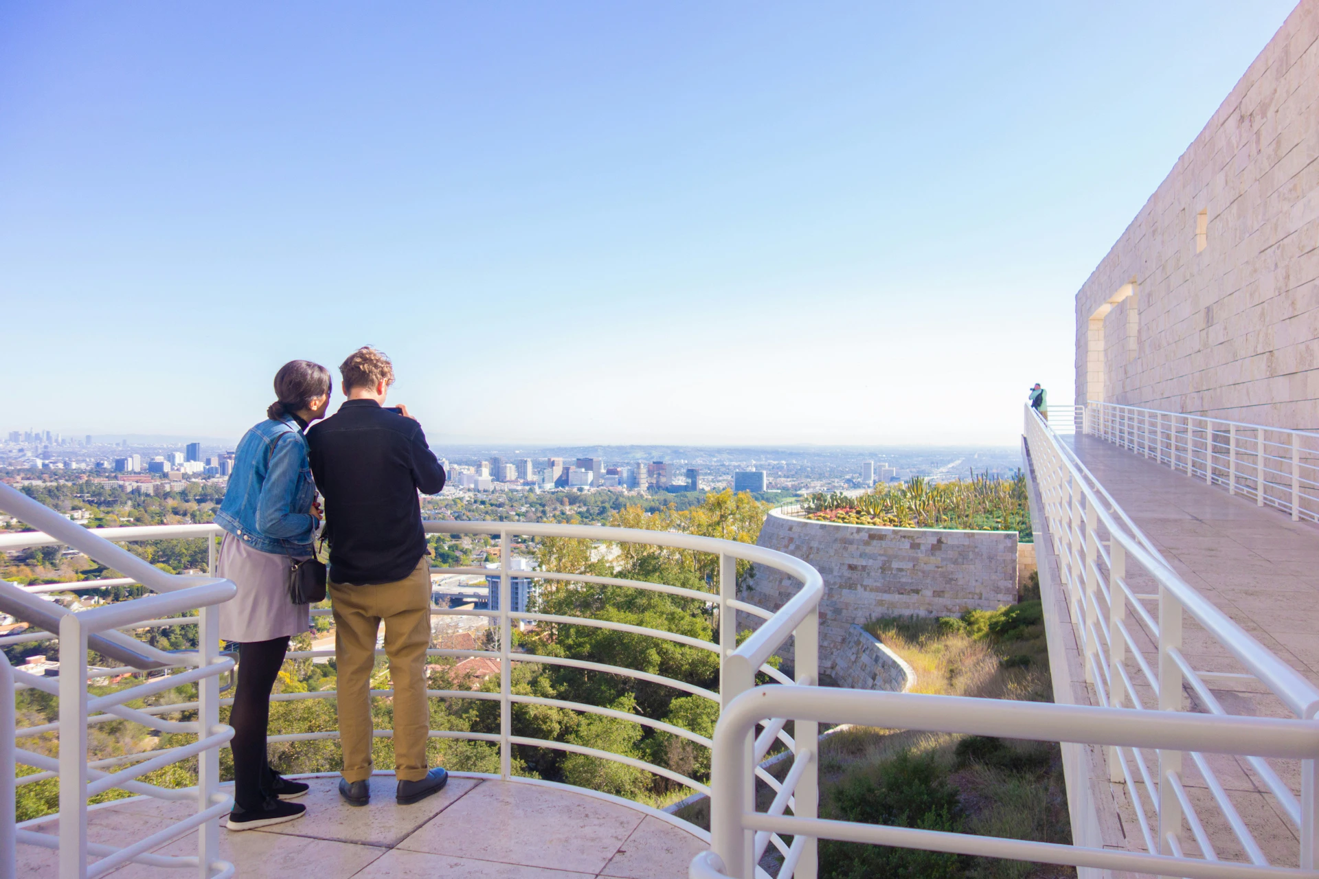 Two people stand on a terrace overlooking a cityscape, with railing and a clear blue sky in the background.