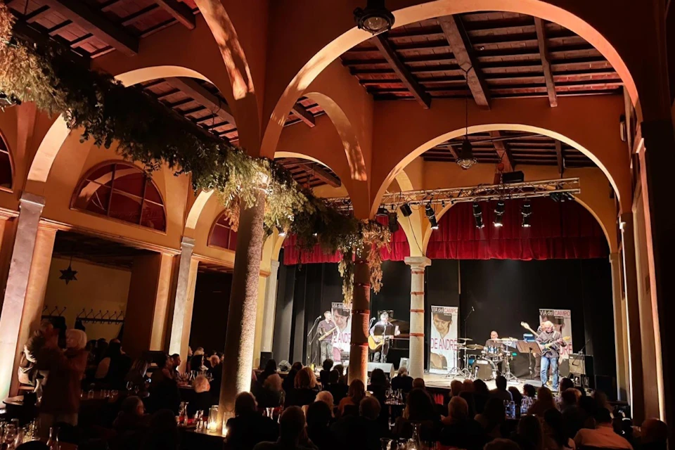 A band performs on a stage under arched ceilings in a dimly lit venue, with an audience seated at tables.