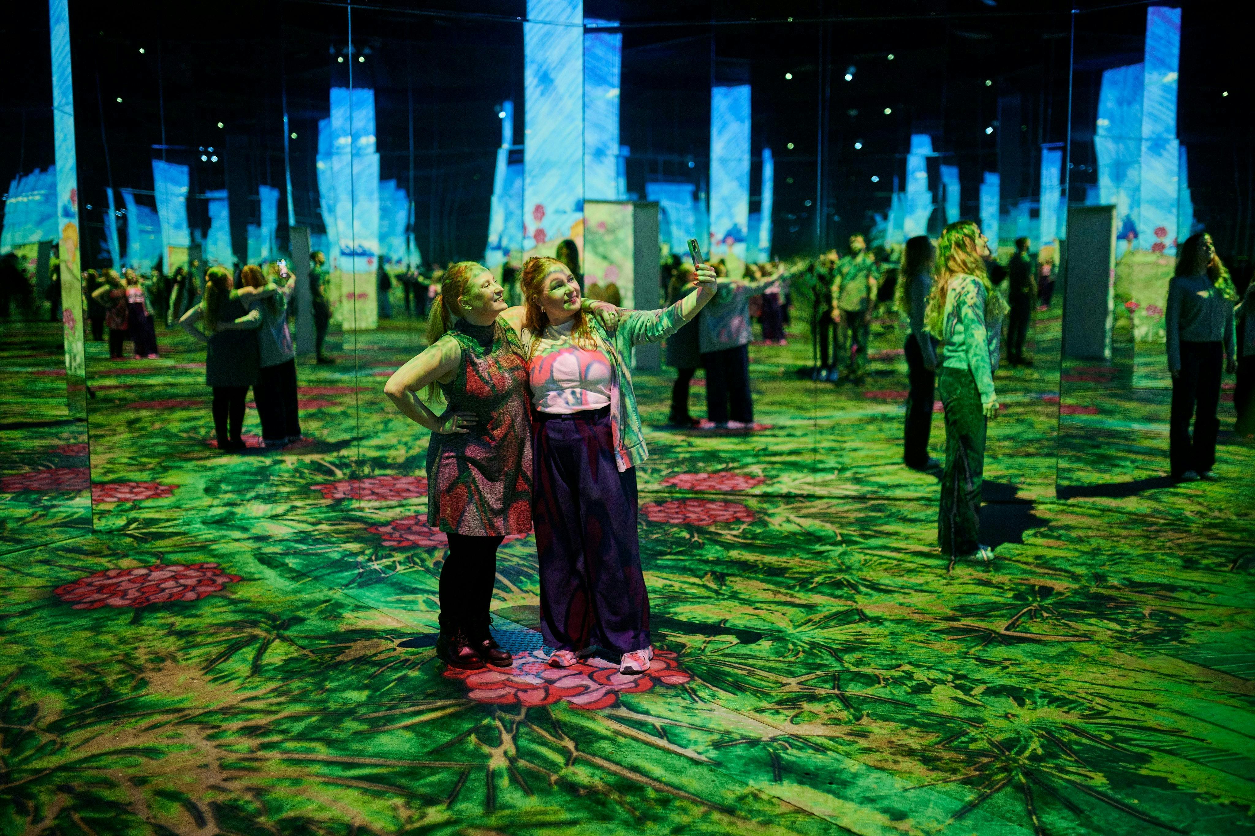 Δύο γυναίκες ποζάρουν για μια selfie σε μια πολύχρωμη, καθηλωτική εικαστική εγκατάσταση με καθρέφτες και φωτιζόμενα σχέδια.