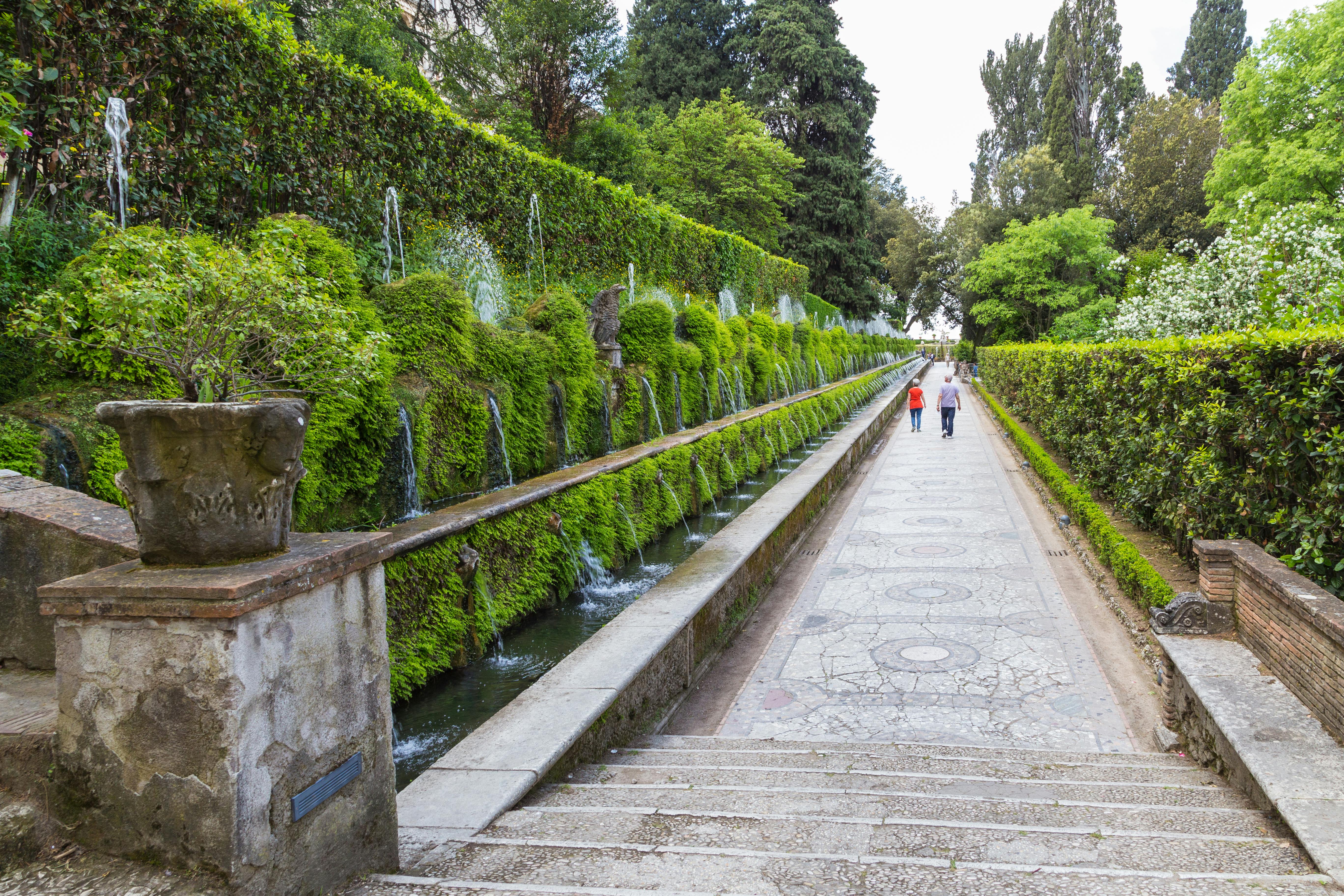 A long, narrow garden path flanked by greenery and fountains, with two people walking away in the distance.