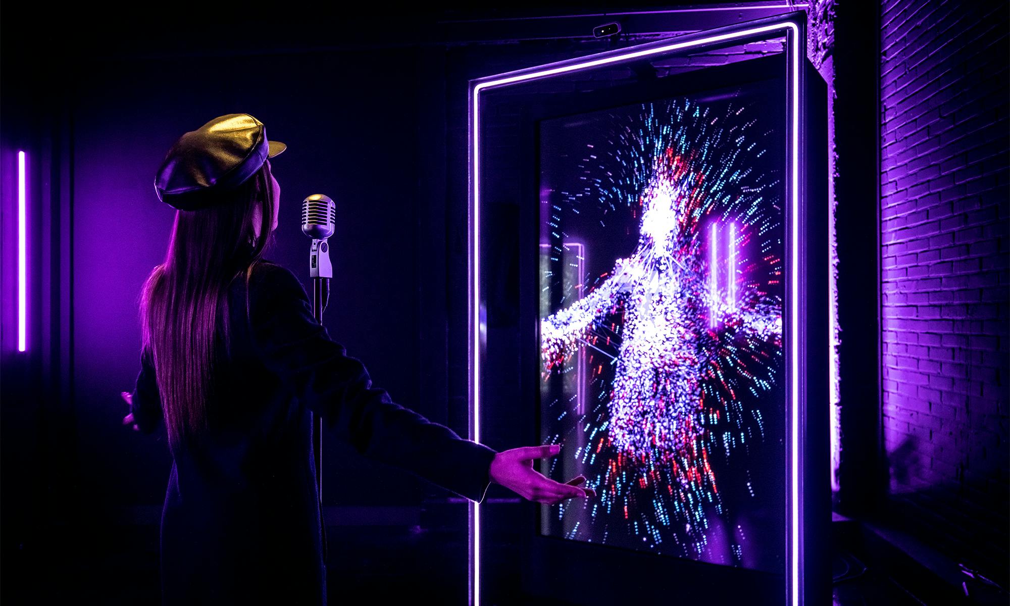 Une personne vêtue d'une tenue et d'un chapeau sombres chante dans un microphone rétro. Un écran numérique vibrant affiche une figure abstraite et colorée.