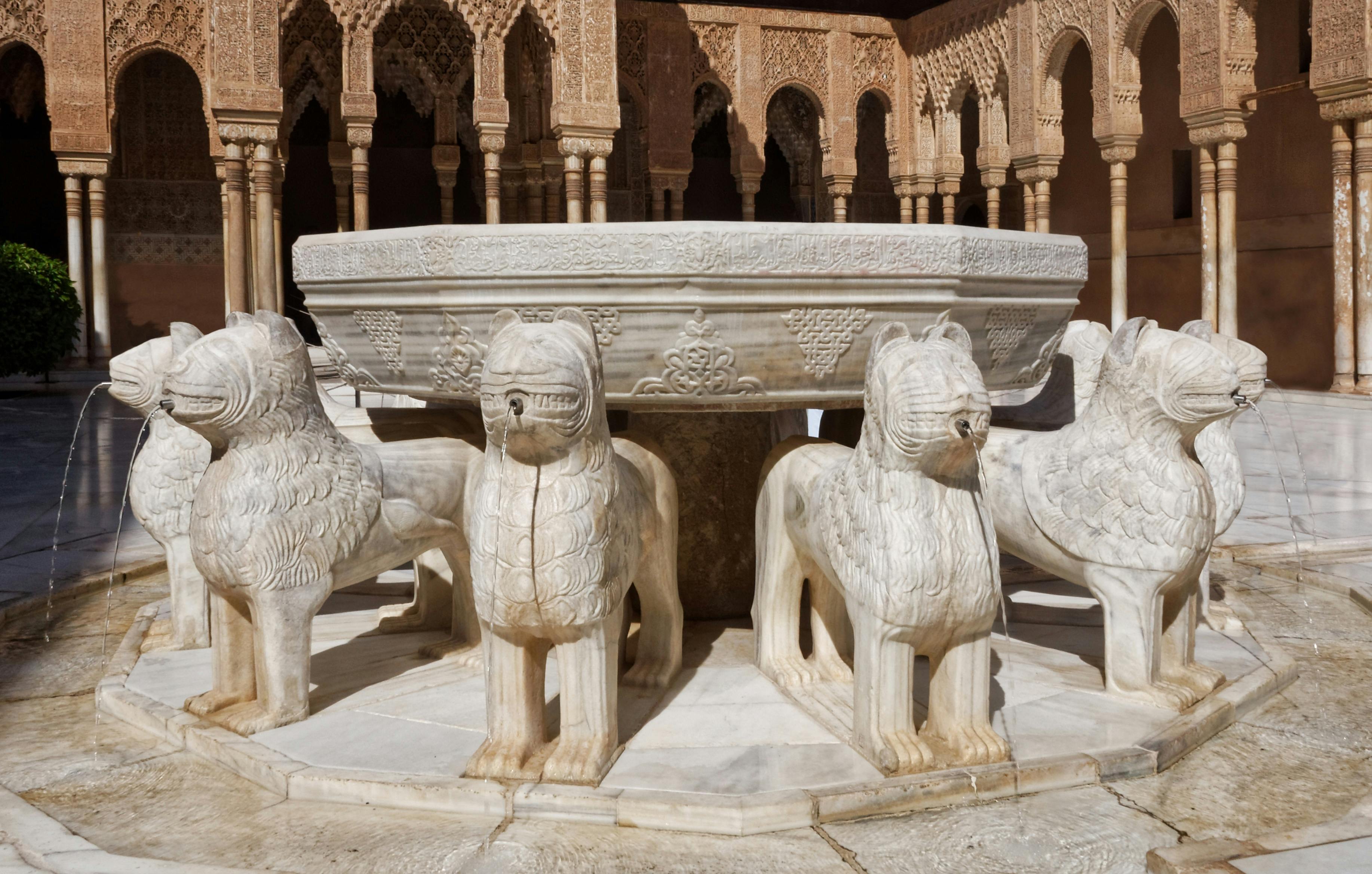 Stone fountain adorned with twelve lion statues in a courtyard with ornate, arched columns in the background.
