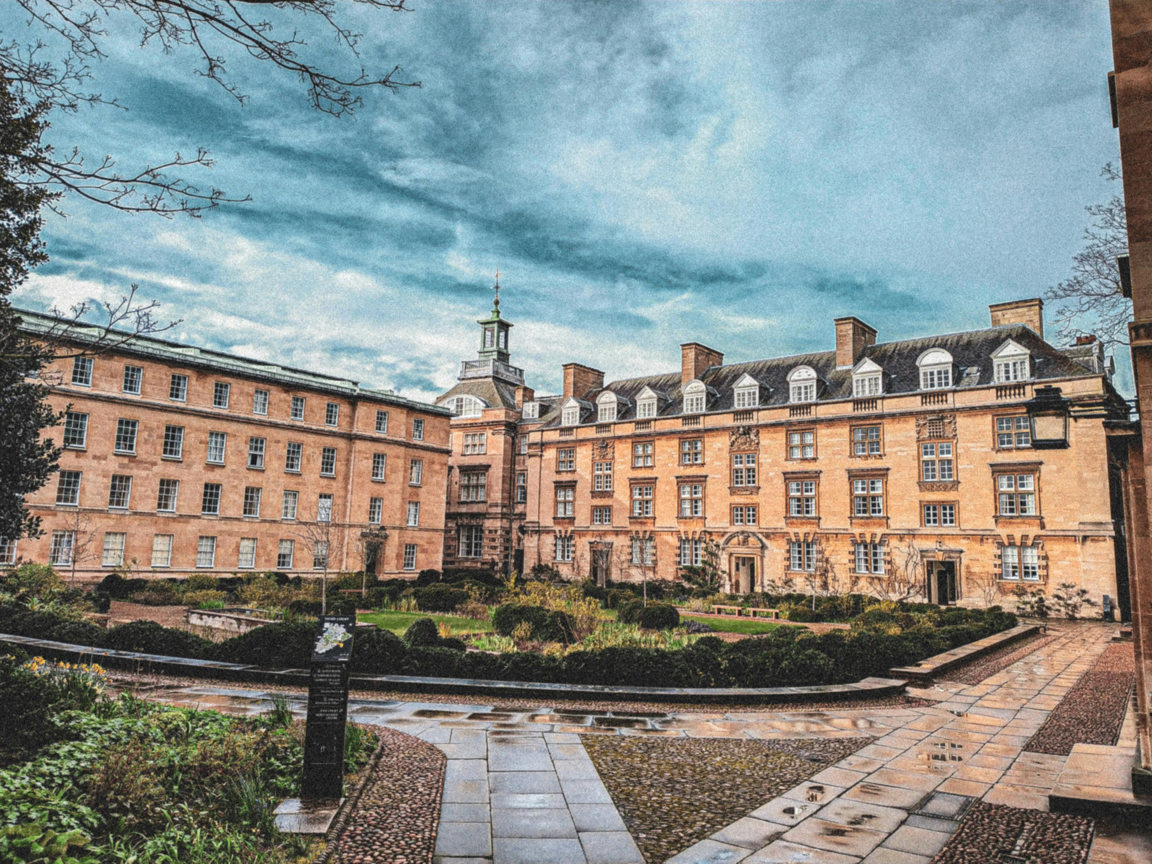 Historic brick building with a central clock tower, surrounded by a courtyard with pathways and hedges under a cloudy sky.