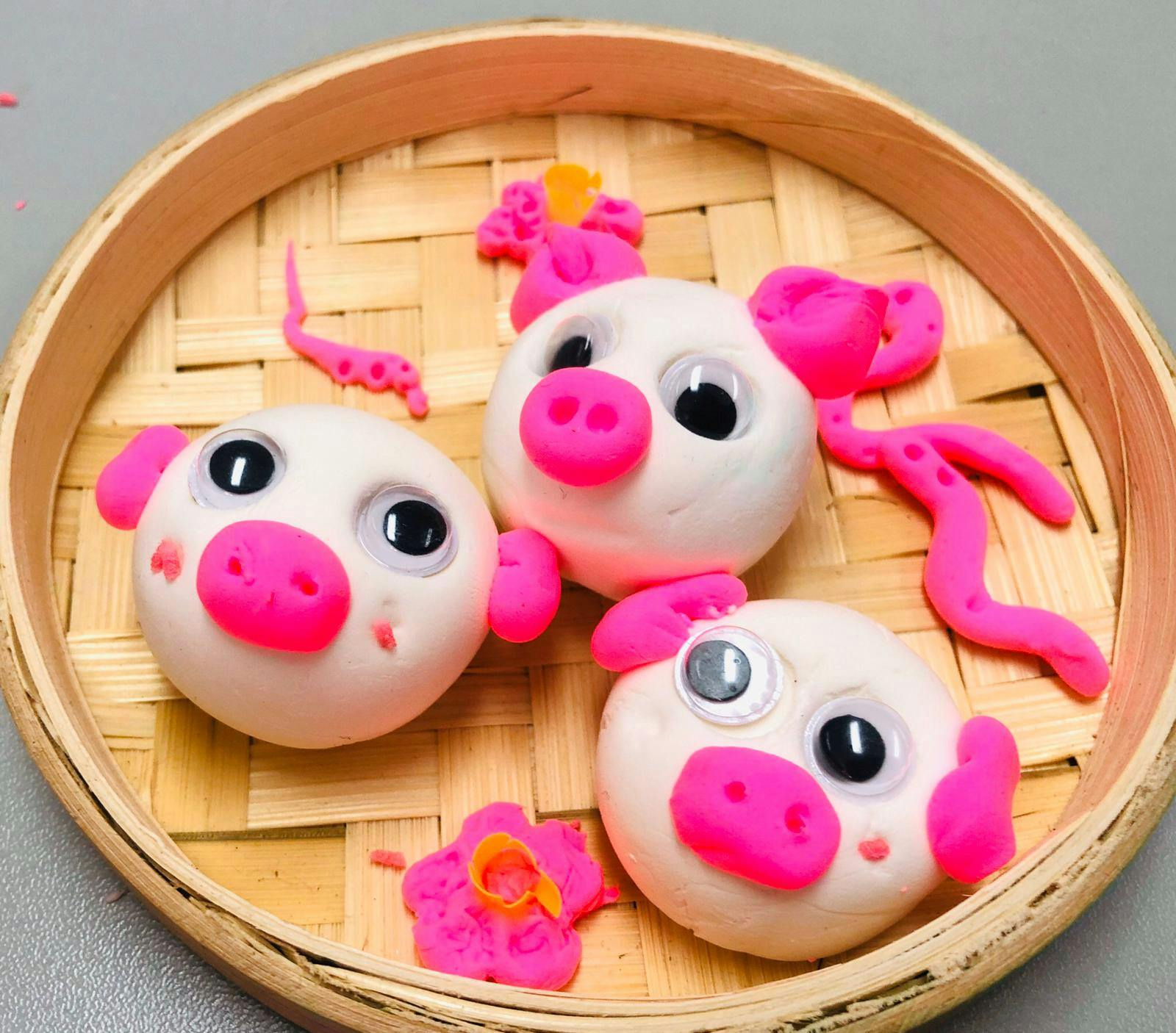 Three pig-shaped buns with pink details and googly eyes in a bamboo steamer, surrounded by pink dough pieces.