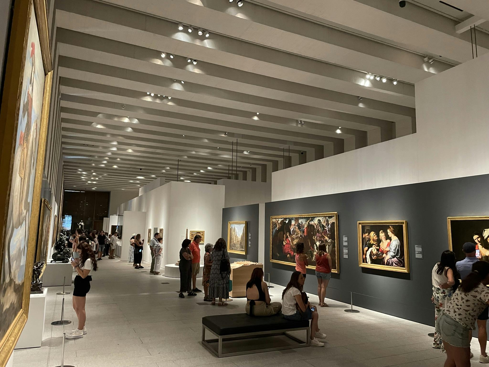 游客在宽敞、光线充足的艺术画廊观赏绘画作品，画廊配有现代化的天花板灯和座椅长凳。