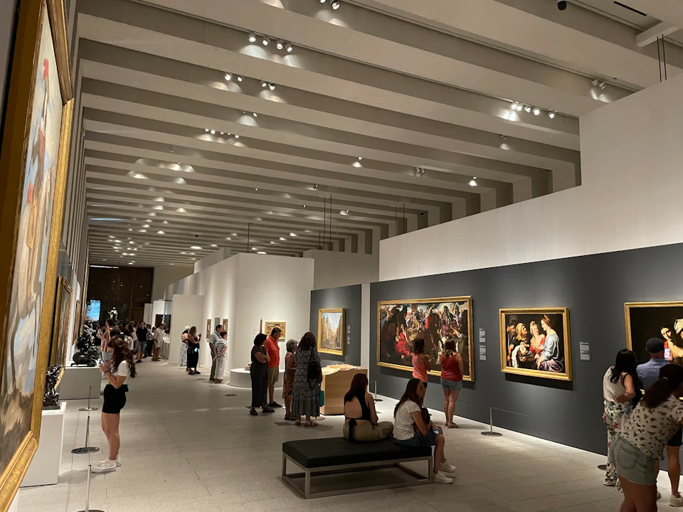 Visitors observing paintings in a spacious, well-lit art gallery with modern ceiling lights and seating benches.