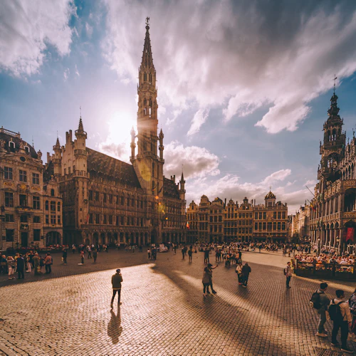 Ett livligt torg med människor, historiska byggnader och ett högt torn under en delvis molnig himmel med solljus som strömmar igenom.