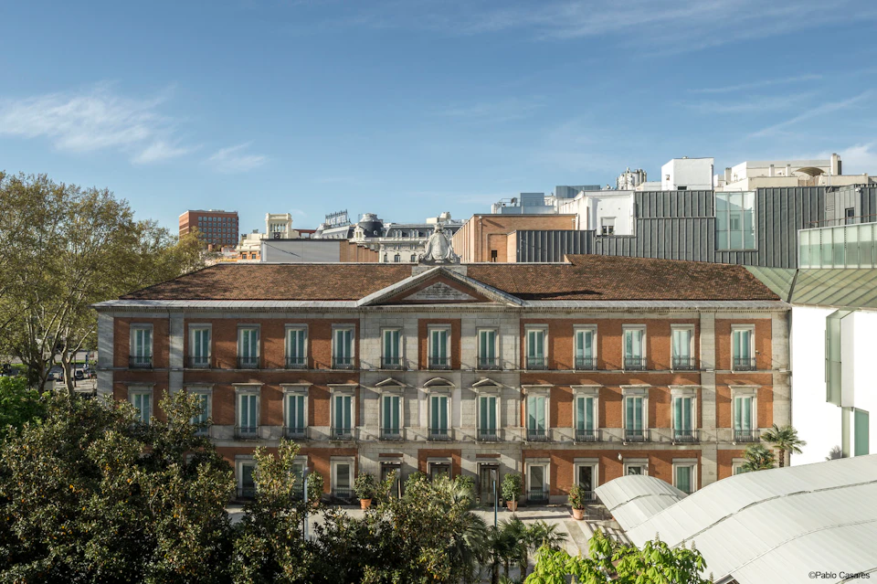 A multi-story red-brick and stone building with numerous windows, surrounded by greenery, set against a cityscape and blue sky.