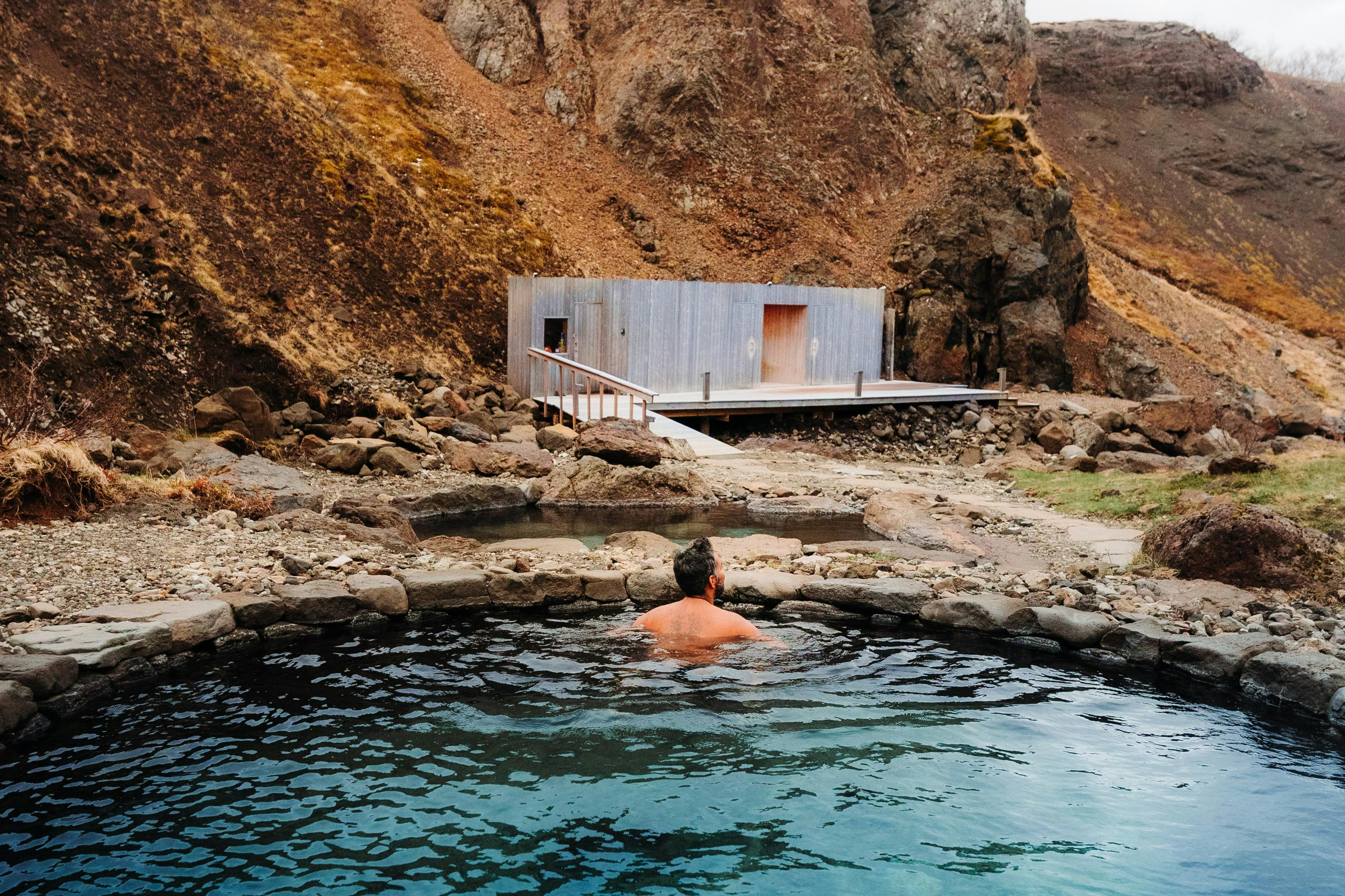 Una persona si rilassa in una piscina di acqua calda rocciosa, con una piccola struttura moderna e una collina rocciosa sullo sfondo.