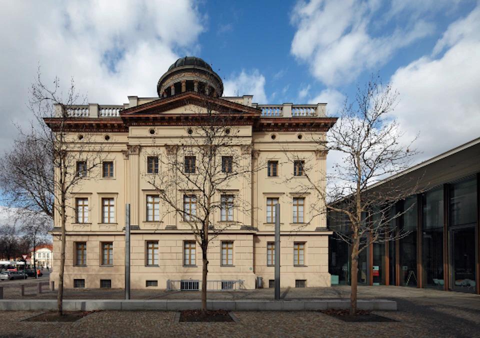 A classical beige building with a dome, large windows, bare trees, and a cobblestone street under a partly cloudy sky.