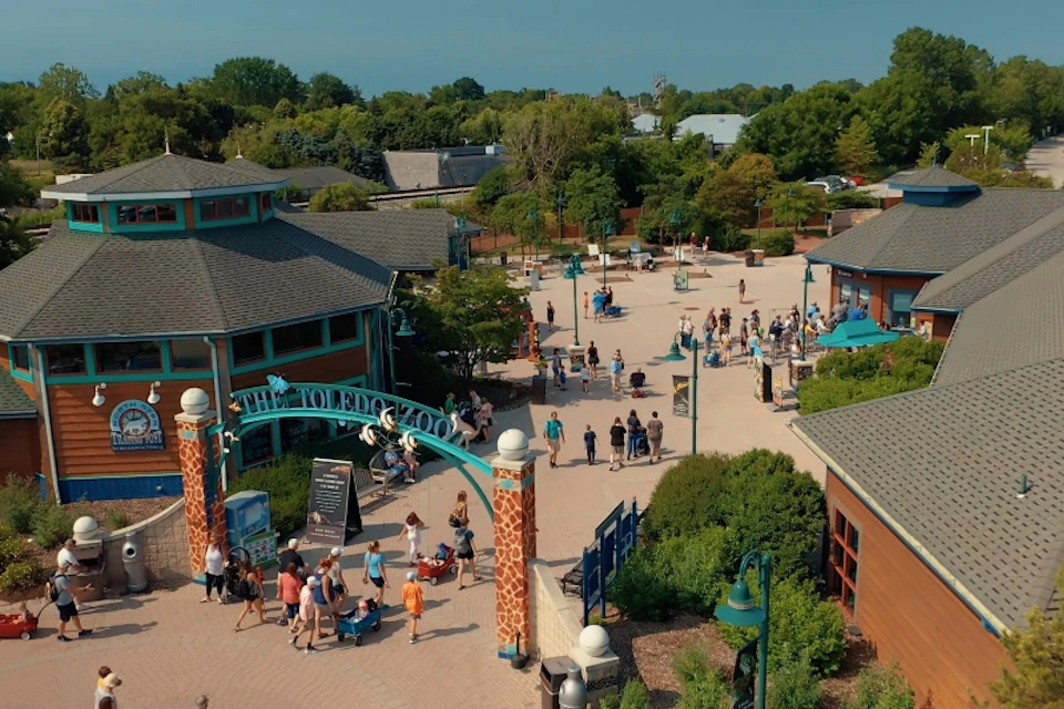 People walking around the entrance of the Toledo Zoo, with nearby buildings and greenery surrounding the plaza.