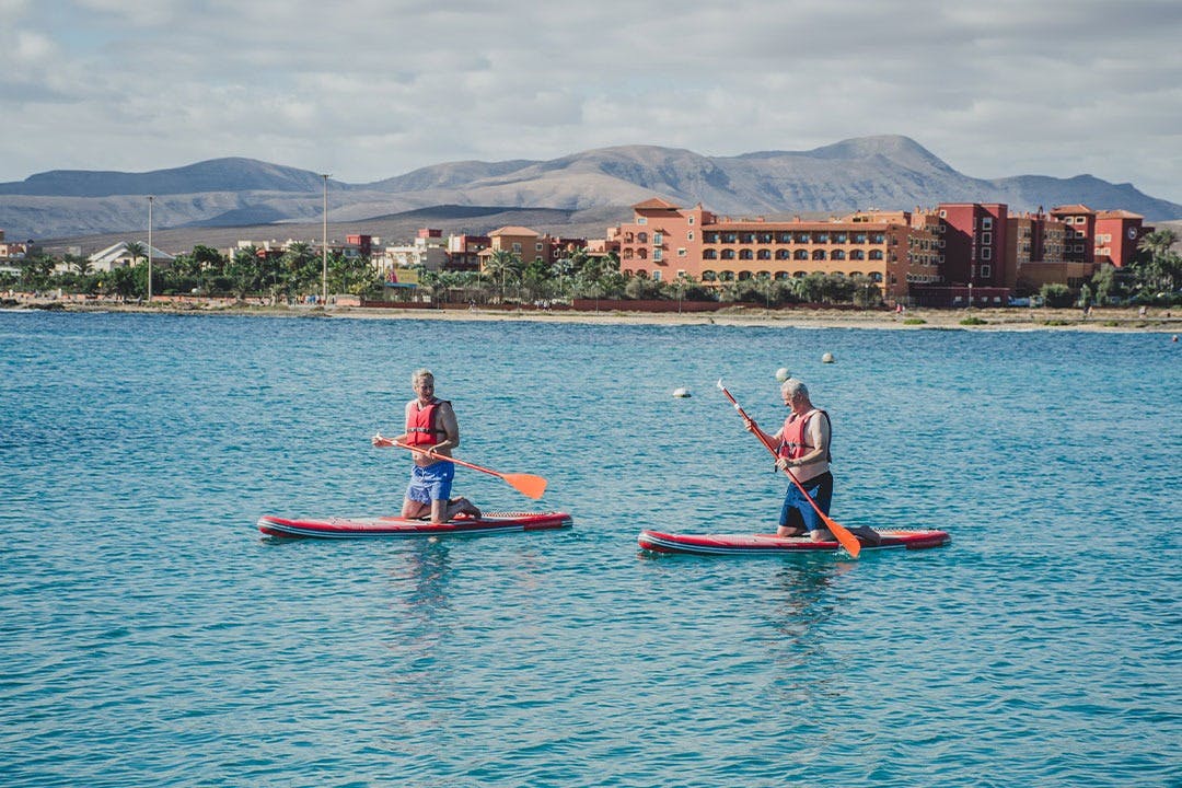 Δύο άνθρωποι κωπηλατούν σε ήρεμα, καταγάλανα νερά με μακρινή θέα σε κτίρια και βουνά κάτω από έναν εν μέρει συννεφιασμένο ουρανό.