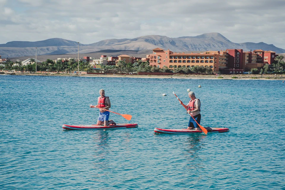 Two people paddleboarding on calm, blue water with a distant view of buildings and mountains under a partly cloudy sky.