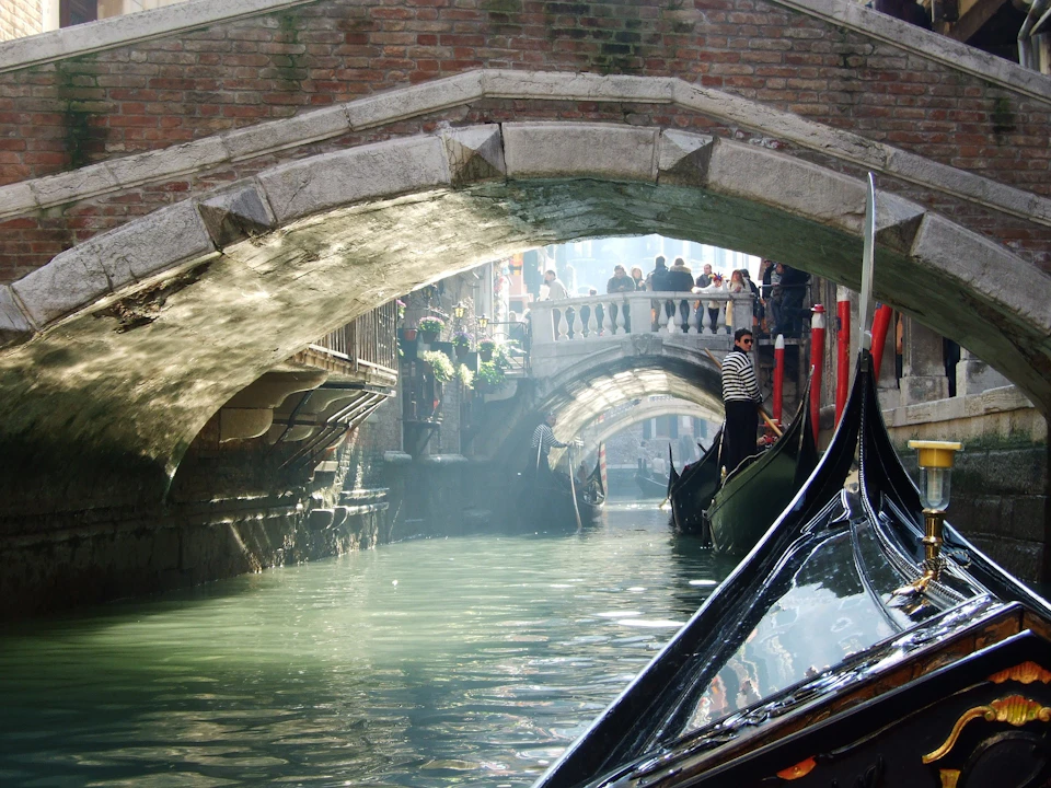 A gondolier navigates a narrow canal under stone bridges with sunlight filtering through, and people observe from above.
