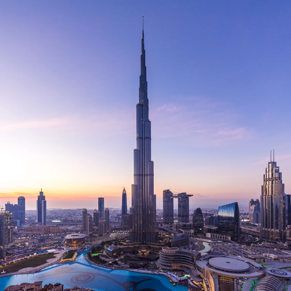 Skyline at sunset featuring a tall skyscraper with surrounding modern buildings and a winding water feature.