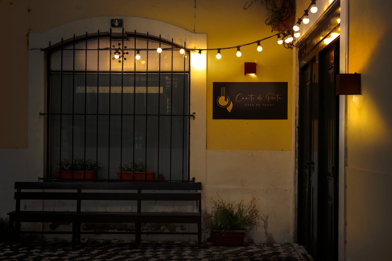 Venkovní kavárenská scéna za soumraku s provázkovými světly a nápisem „Café de Praça“. Lavice a květináče jsou pod oknem.