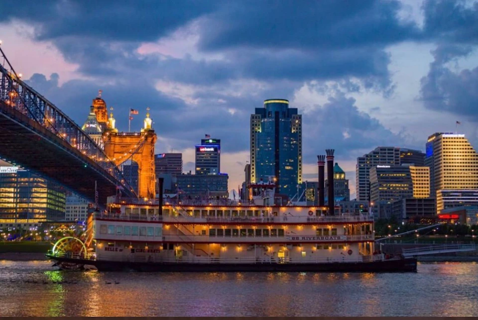 A riverboat illuminated at dusk with a bridge and city skyline in the background, under a partly cloudy sky.