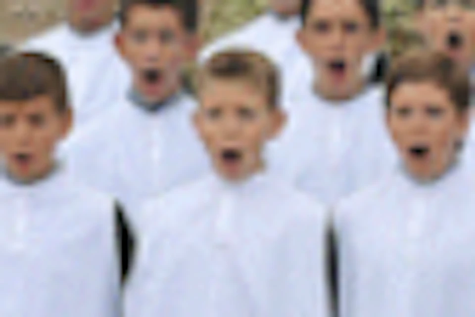 A group of boys in white robes standing in front of a large historic building with a rocky hillside and trees in the background.
