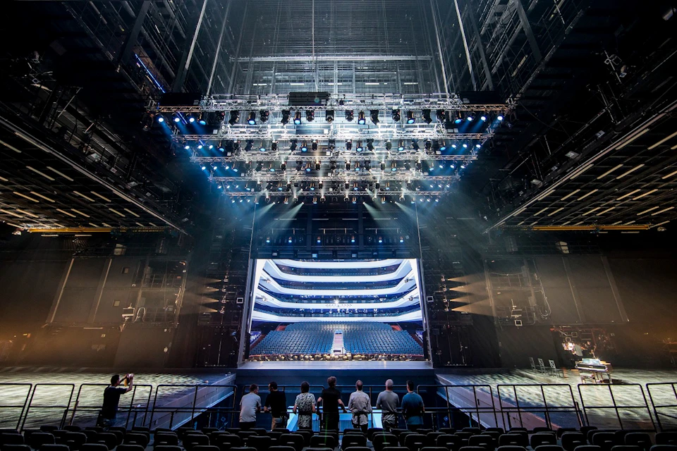 A group of six people stand before an empty stage in a large theater, facing an illuminated set and rows of auditorium seats.