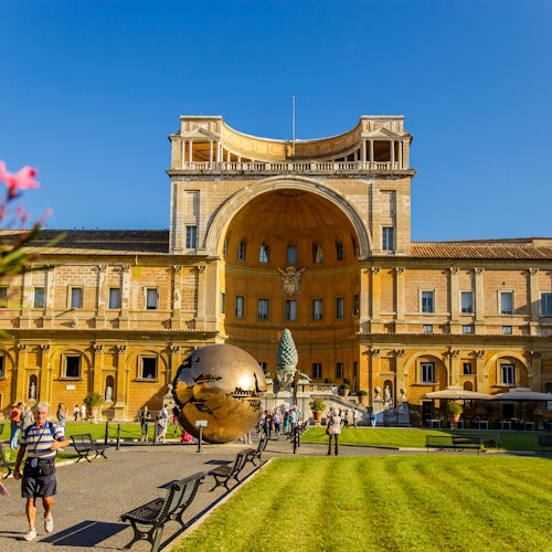 Persone che camminano in un cortile con una grande scultura in bronzo a forma di sfera, circondata da edifici storici e prati verdi; il cielo è azzurro.