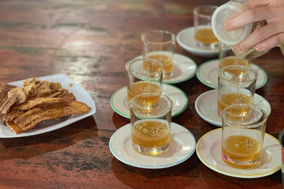 Several small glasses of beer on saucers are being filled on a table, with a plate of fried snacks to the side.