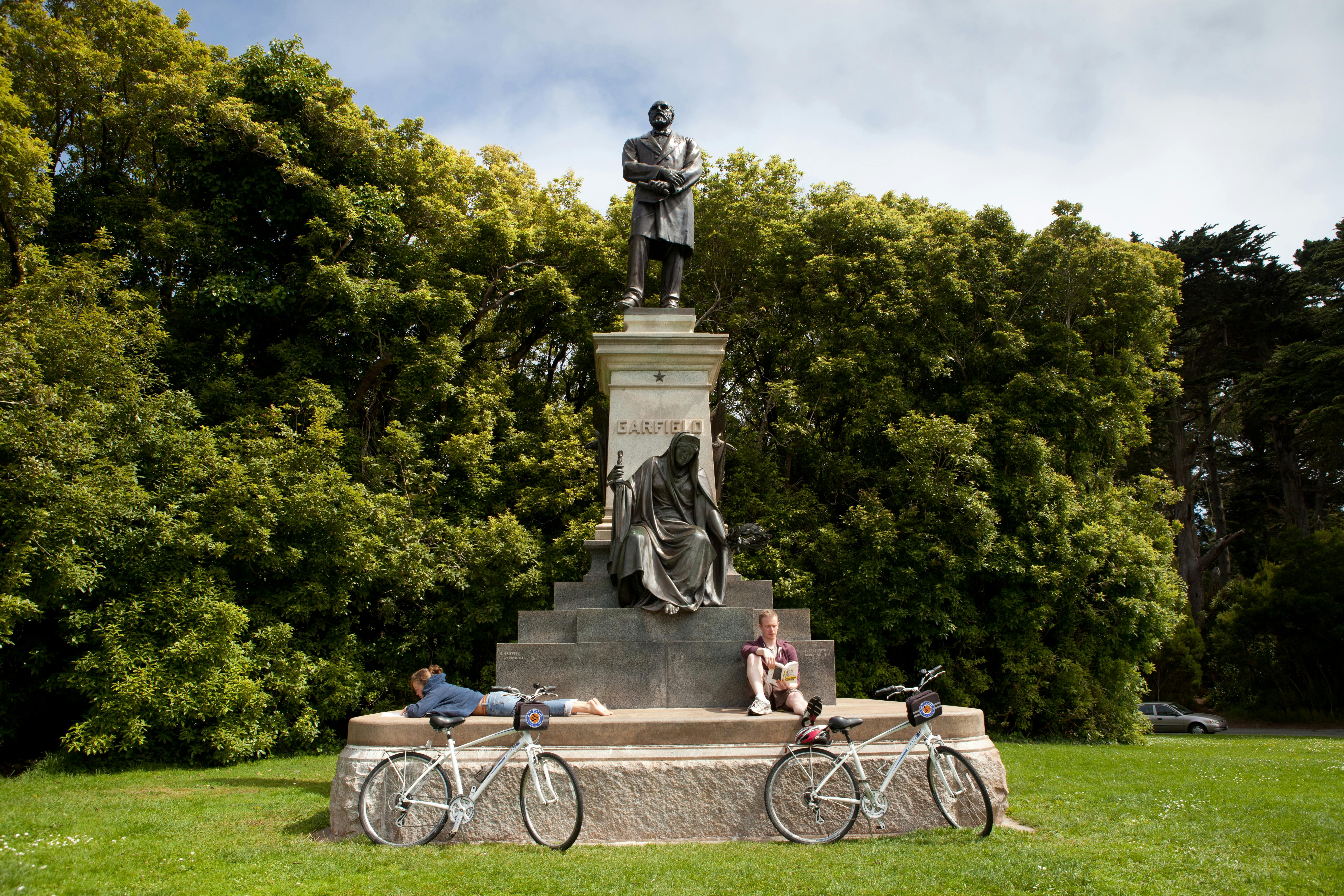 Two people rest near a statue with the name "Garfield" on it. Two bicycles are leaned against the statue's base. Trees are in the background.