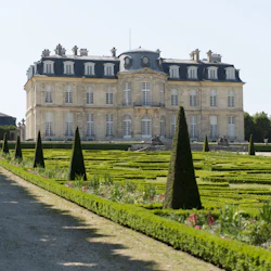 Formal garden with manicured hedges and topiary trees in front of a large, ornate mansion with multiple windows and a steep roof.