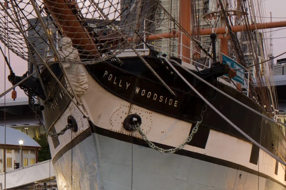 The bow of a historic ship named Polly Woodside, with rigging and chains visible, docked at a maritime display area.