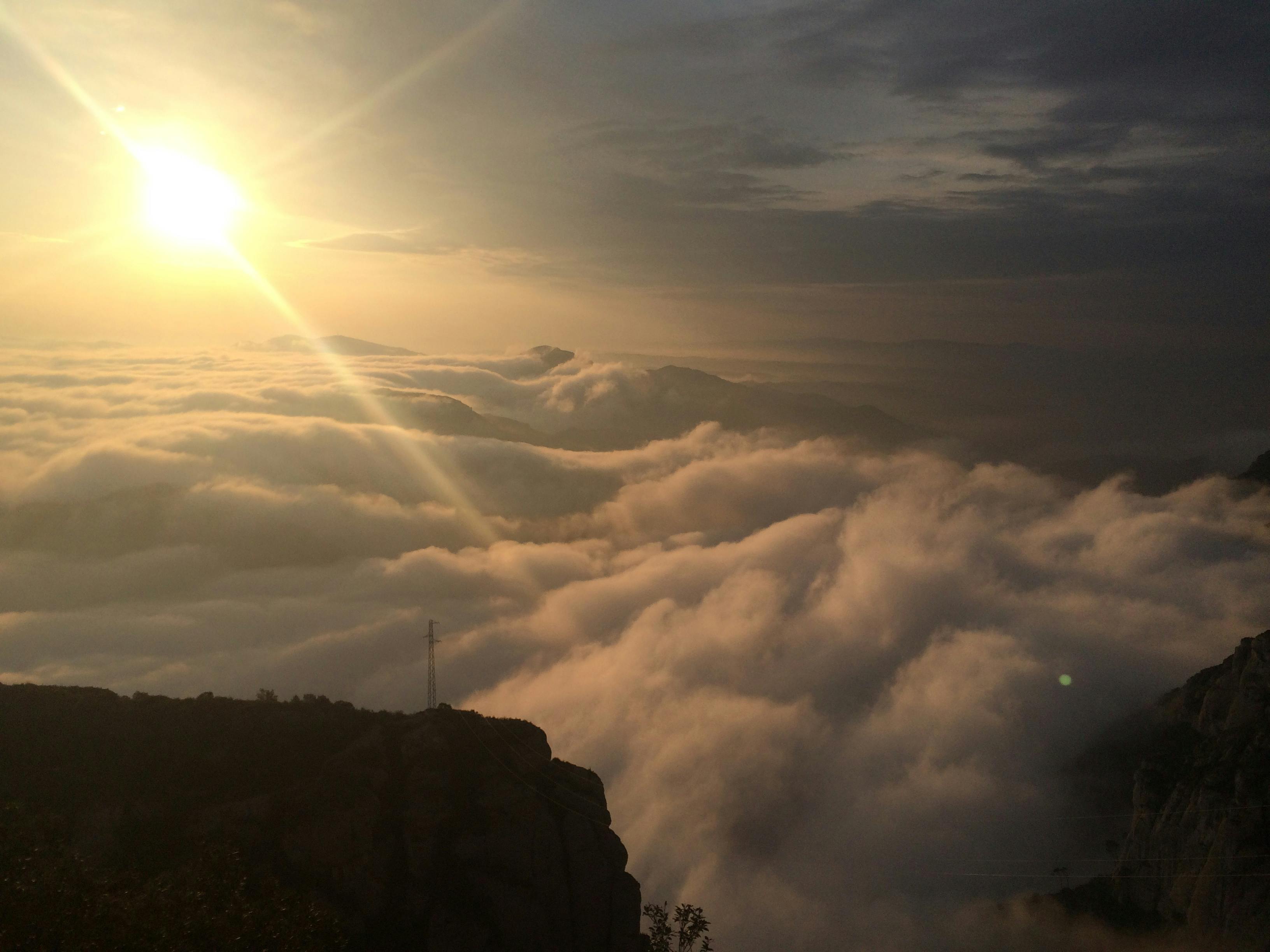 The image shows a sunrise or sunset over a mountain range with dense clouds below, creating a serene and misty atmosphere.