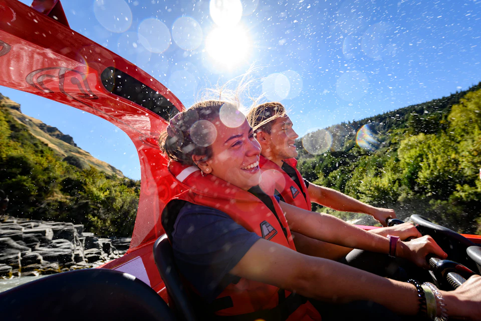 Two people wearing red life jackets on a boat ride, smiling, with water splashes and bright sunlight in a natural setting.