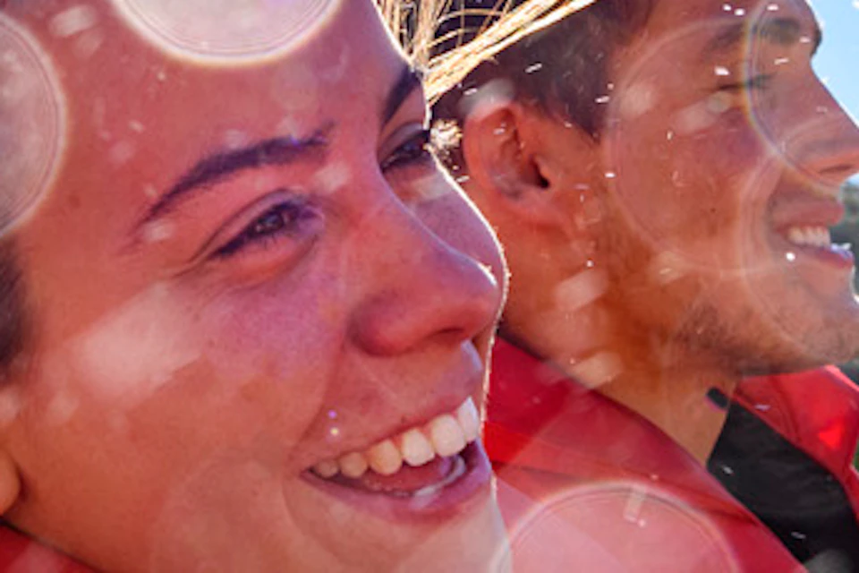 Two people wearing red life jackets on a boat ride, smiling, with water splashes and bright sunlight in a natural setting.