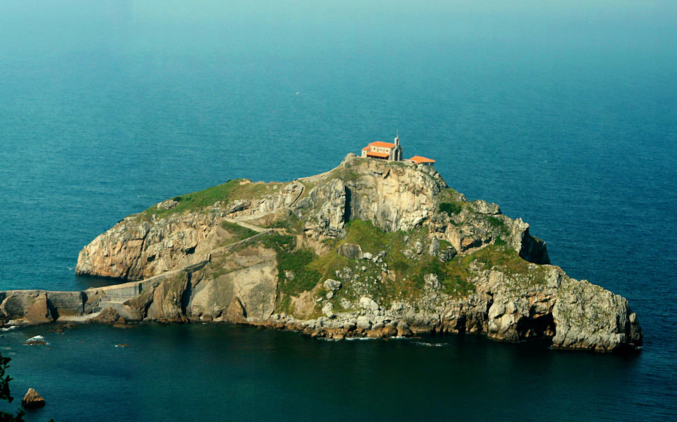 A small chapel with an orange roof atop a rocky island surrounded by the sea, with a winding path leading up to it.