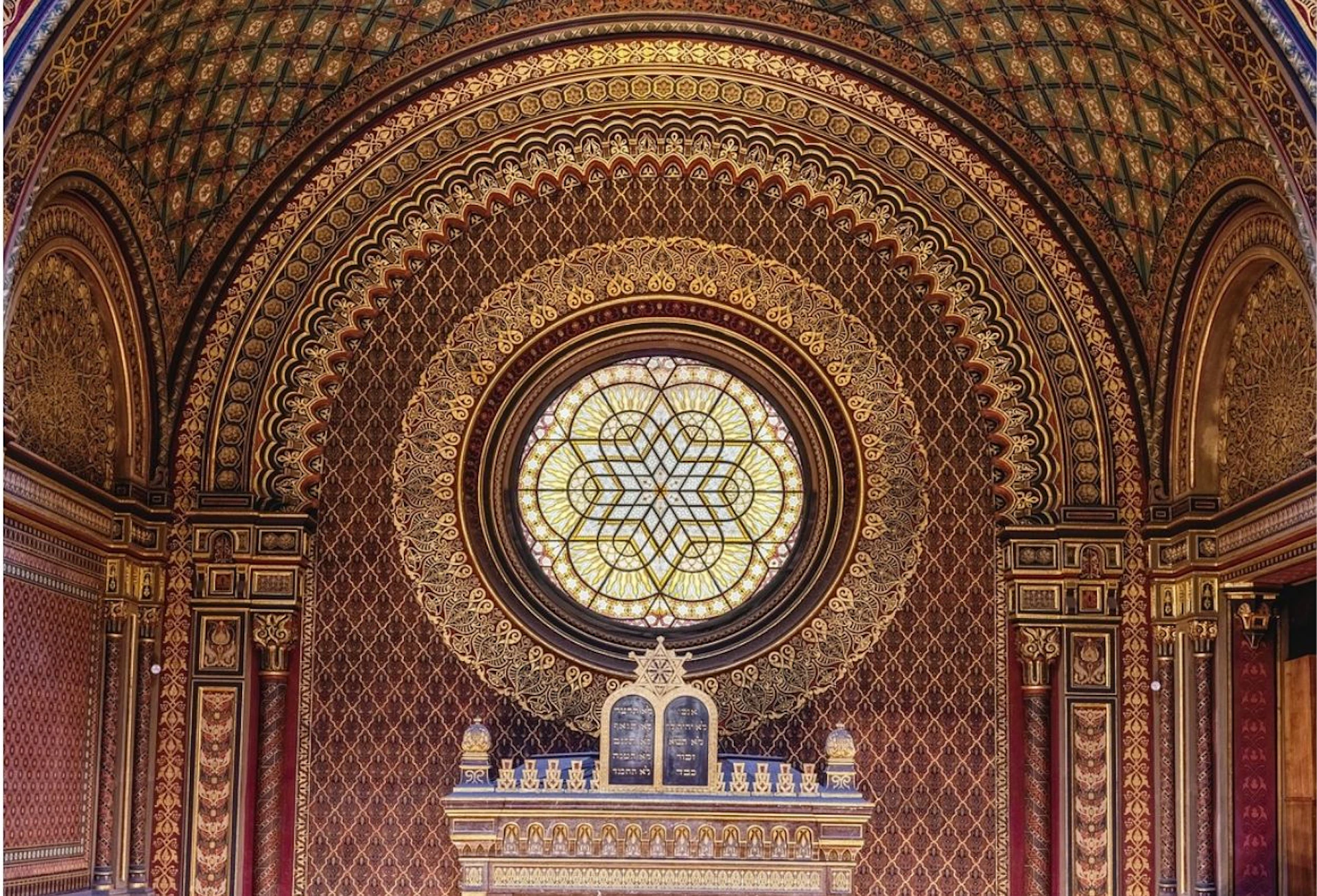 Elaborately decorated arched wall with a circular stained glass window and intricate patterns in gold, red, and blue.