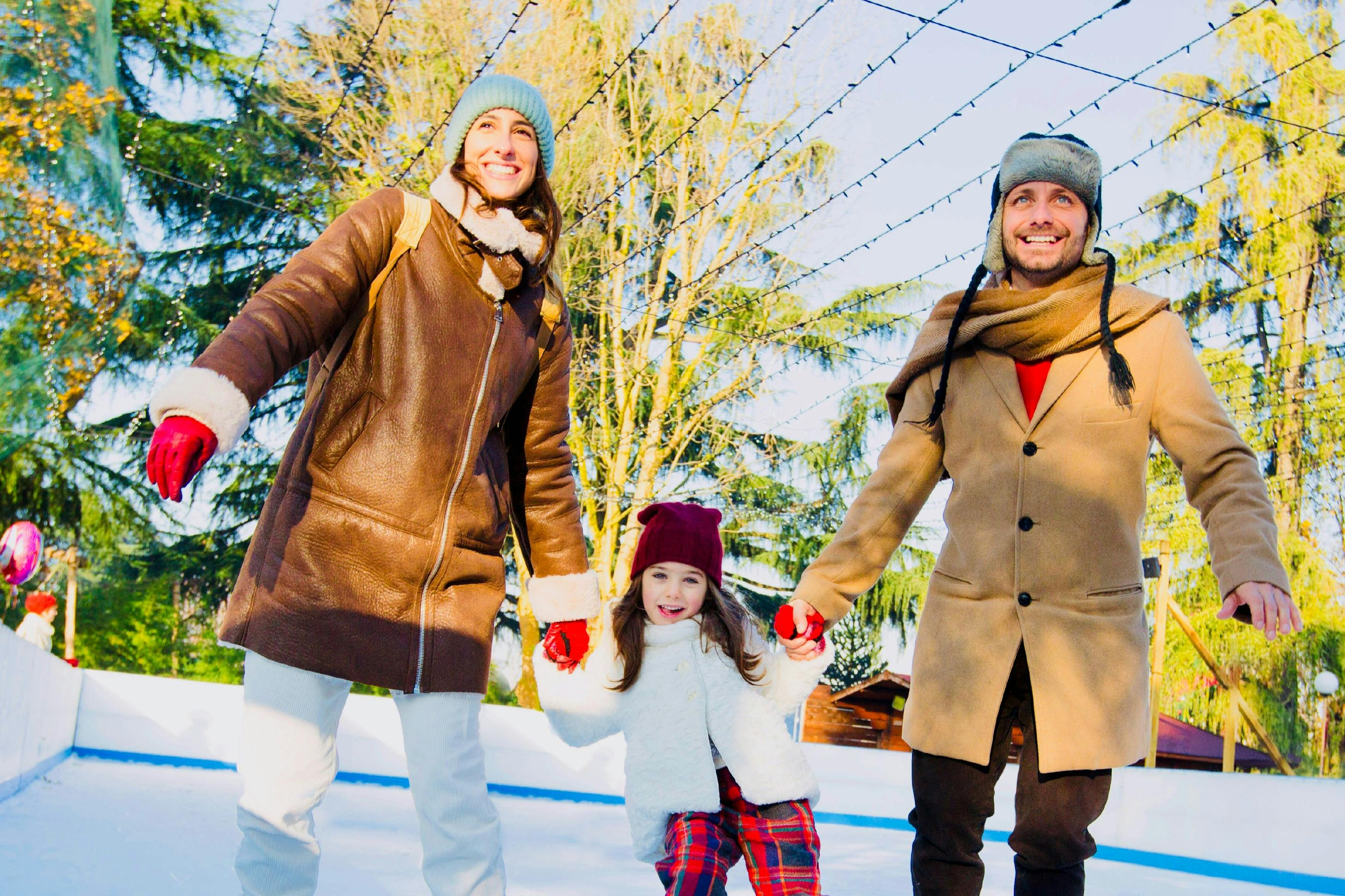 Μια χαμογελαστή οικογένεια κρατώντας τα χέρια, ντυμένη με χειμωνιάτικα ρούχα, πατινάζ στον πάγο σε εξωτερικούς χώρους με δέντρα και φωτάκια στο παρασκήνιο.