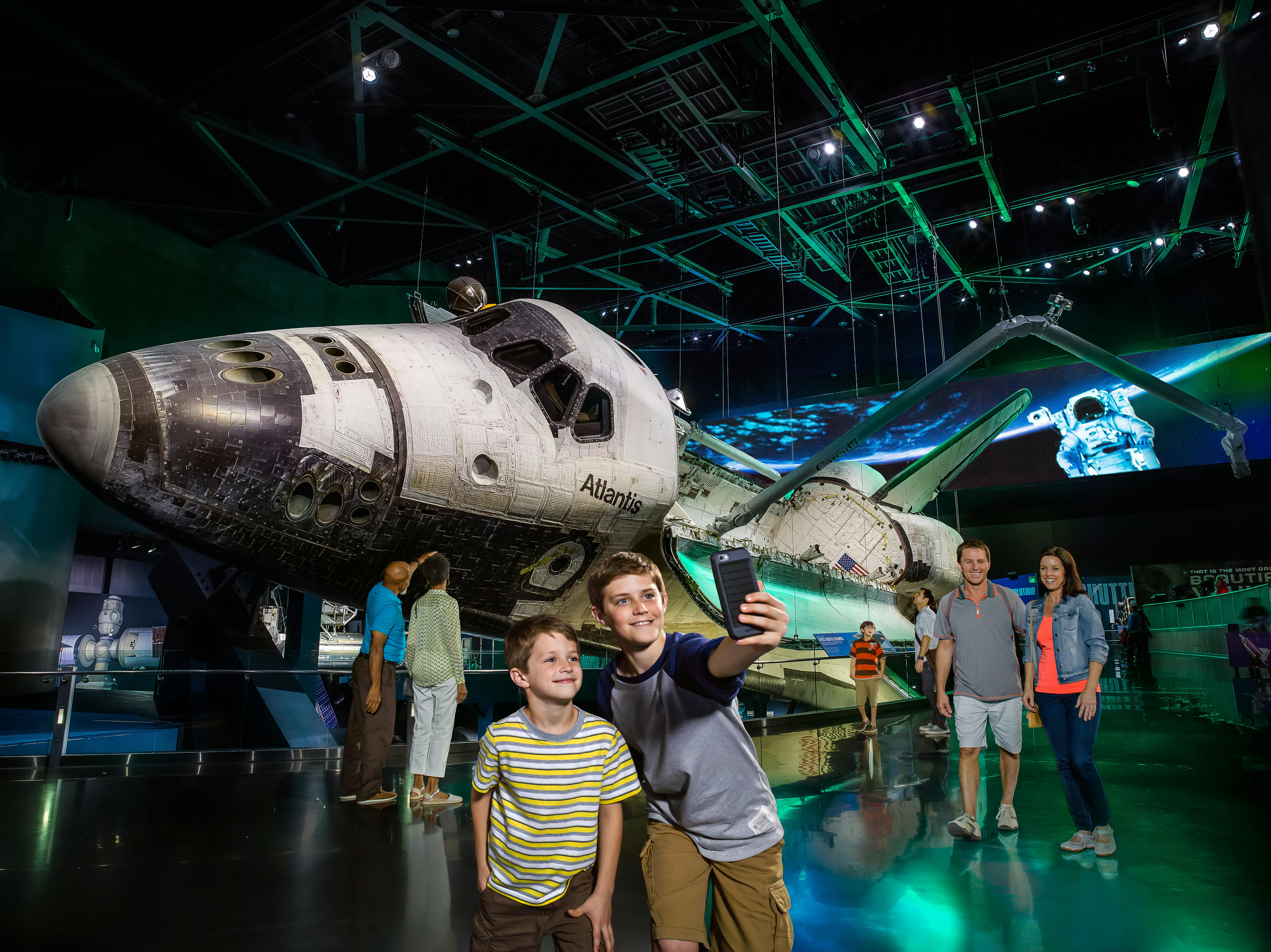 Two boys take a selfie in front of the space shuttle Atlantis, displayed in a museum with dramatic lighting.