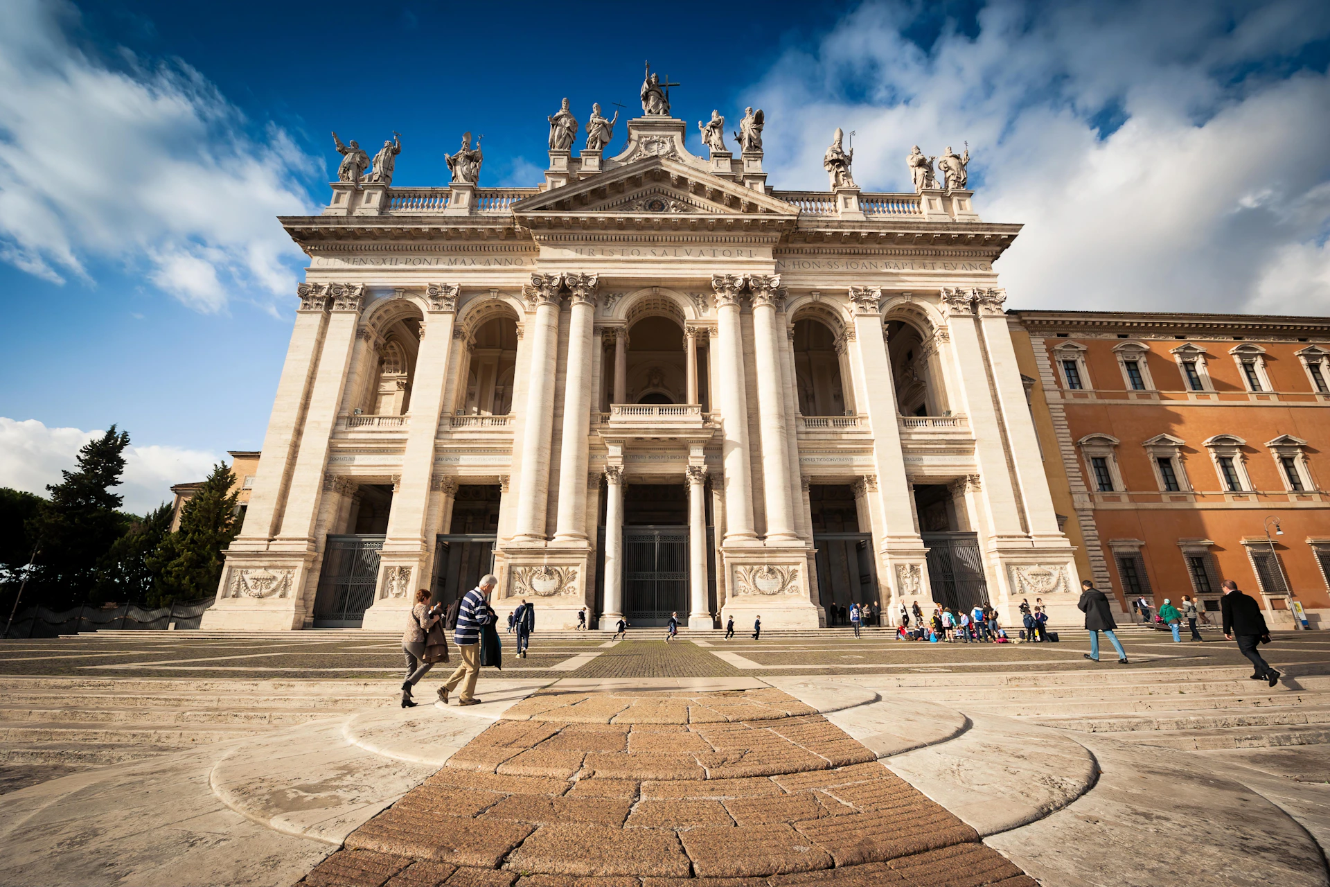 A grand historical building with ornate columns and statues, people walking and gathering in the foreground.