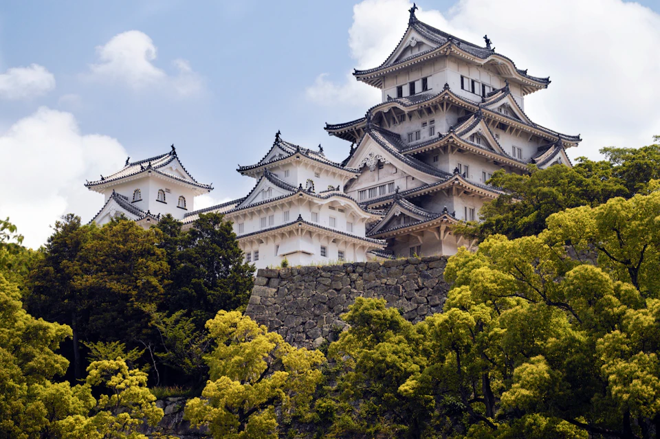 A traditional Japanese castle with white walls and tiled roofs surrounded by green trees under a blue sky with clouds.