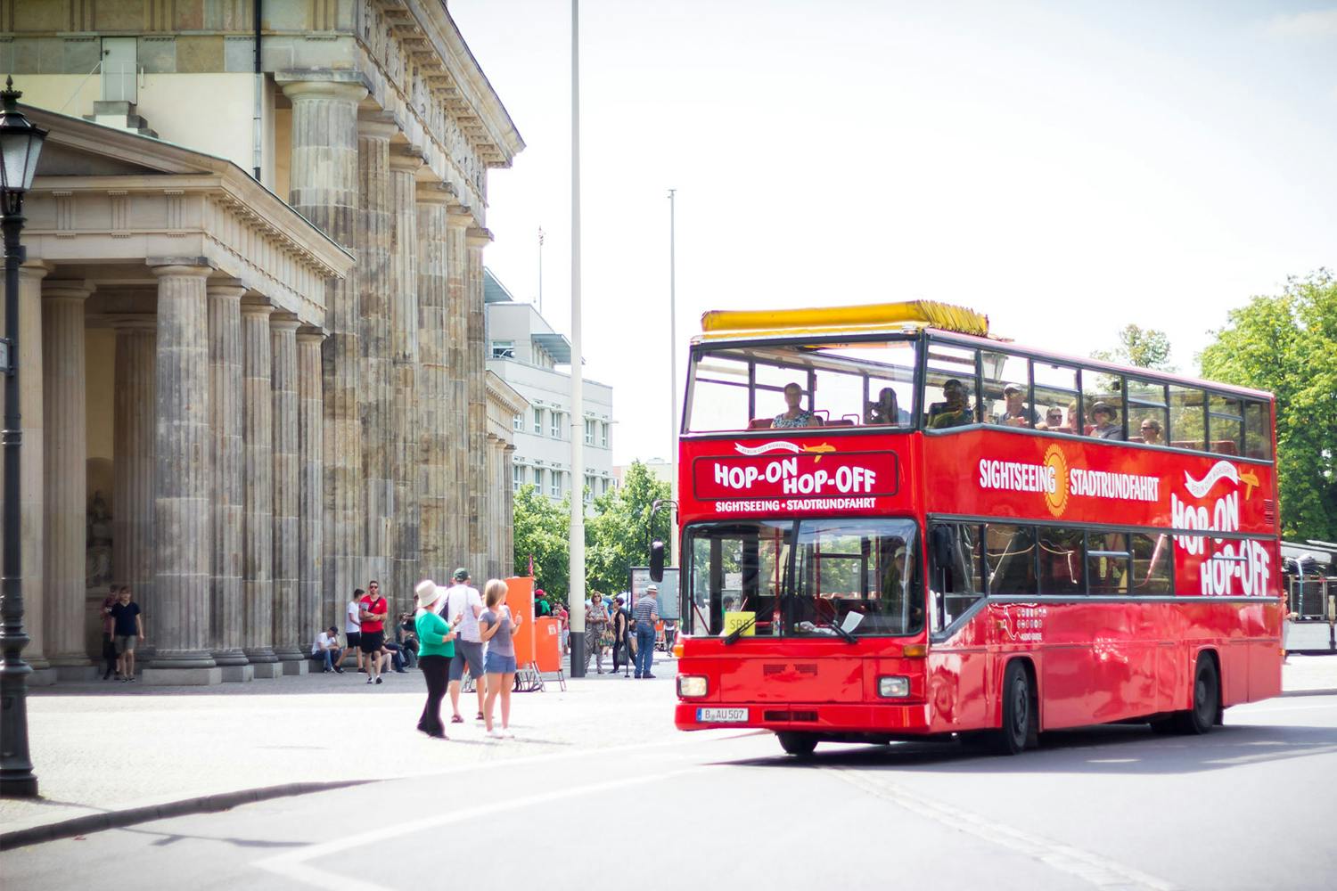 Un bus touristique rouge à impériale portant la mention "Bus Hop-on Hop-off". Des personnes se tiennent près d'un bâtiment historique.