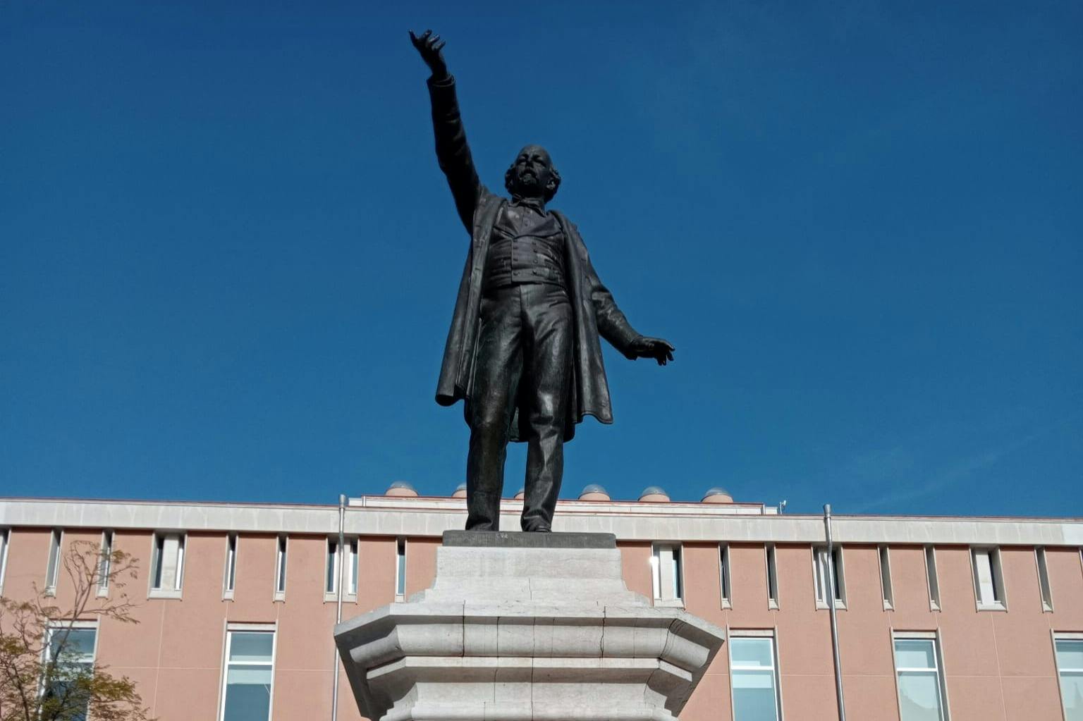 Χάλκινο άγαλμα ενός άνδρα με υψωμένο το χέρι, τοποθετημένο σε πέτρινο βάθρο μπροστά από ένα σύγχρονο κτίριο με φόντο έναν καταγάλανο ουρανό.