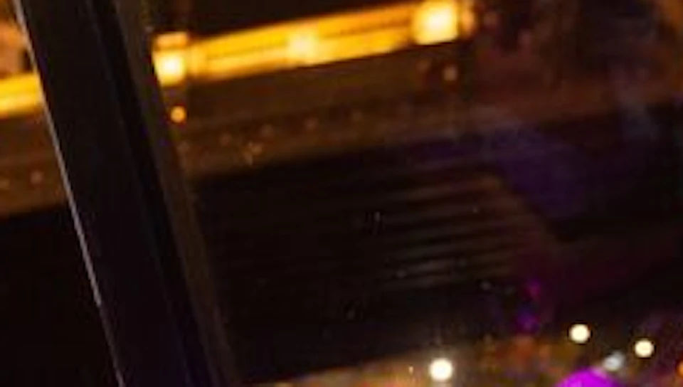A woman in a floral dress toasts with a glass of champagne at a candlelit table by a river at night, with a lit bridge in the background.