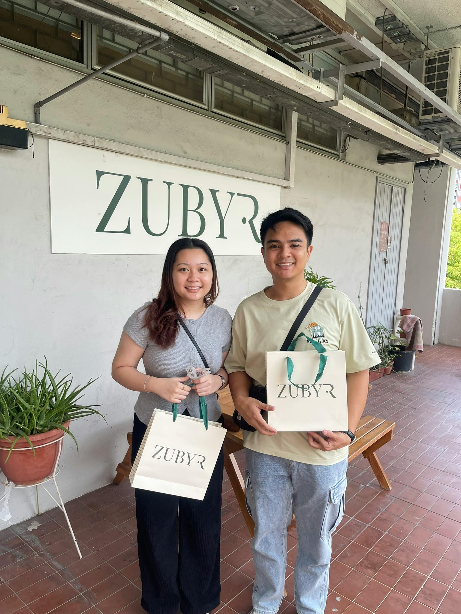 Two people smiling while holding Zubyr-branded shopping bags outside a store with the Zubyr sign visible.