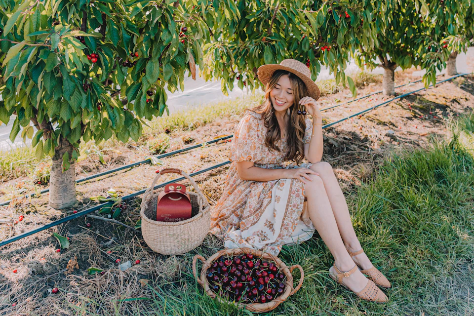 A woman in a floral dress and straw hat sitting on grass near a cherry tree, with baskets of cherries beside her.