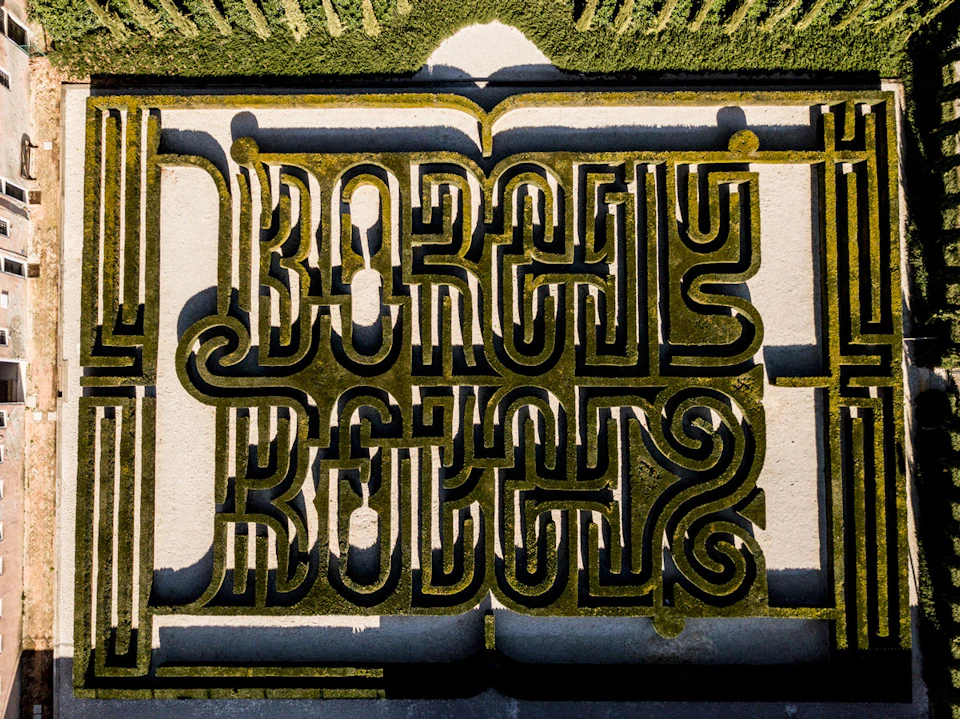 Aerial view of a maze with intricate hedge patterns spelling out "BORGES" symmetrically in green and beige tones.