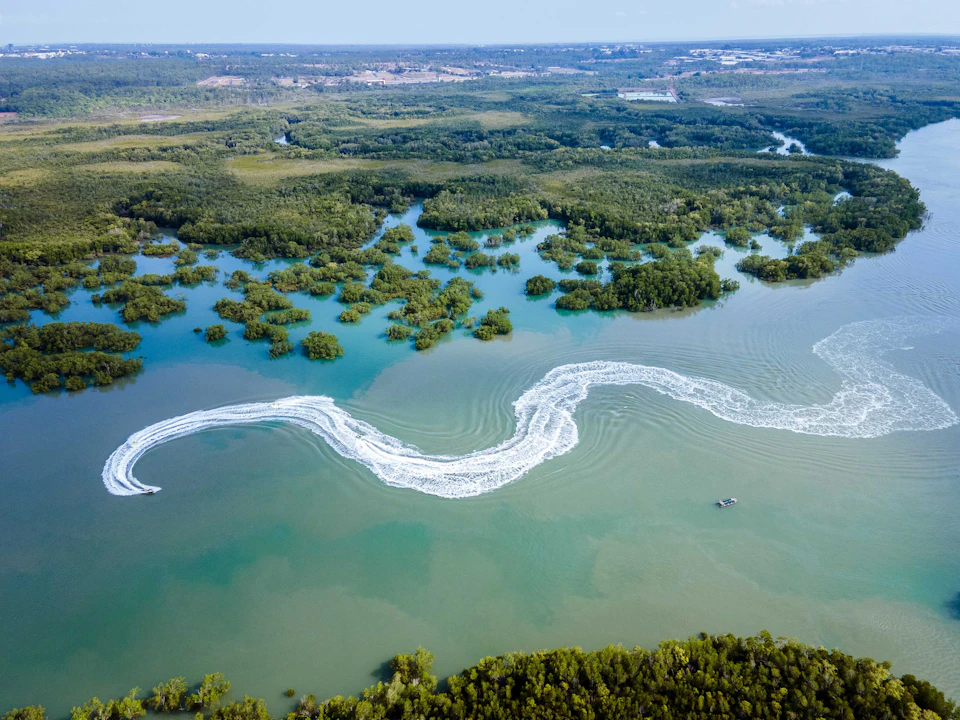 Aerial view of winding river through dense greenery, showing boat wake patterns on the water.