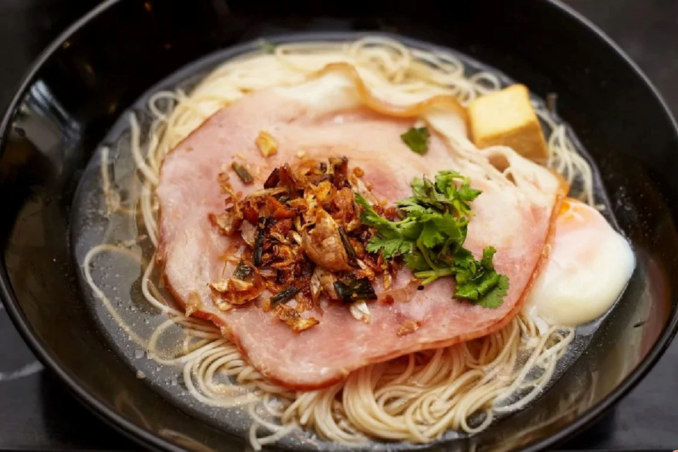 A bowl of noodle soup topped with a slice of ham, fried onions, cilantro, and a piece of tofu.