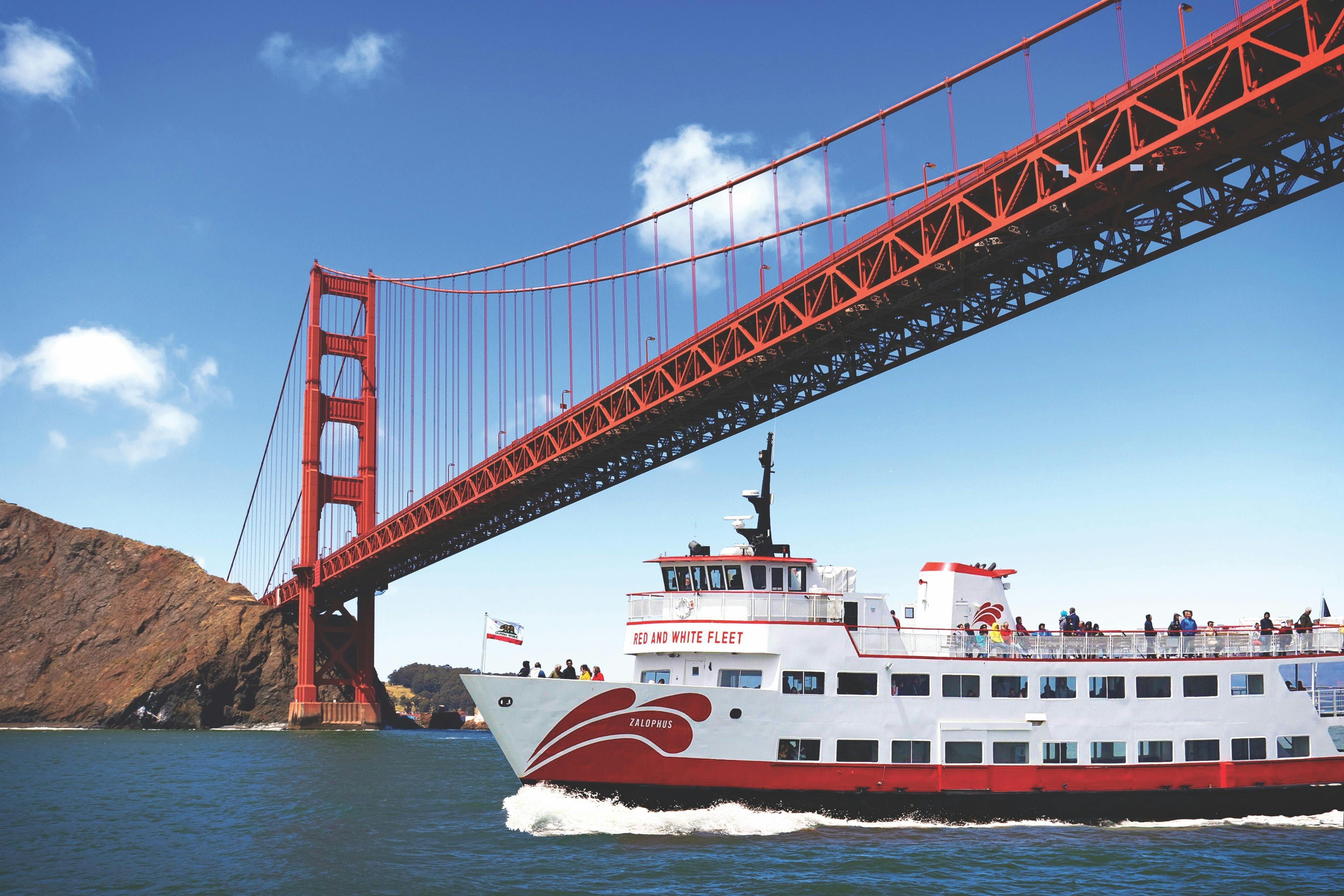 A white and red tour ferry passes under the Golden Gate Bridge on a sunny day.