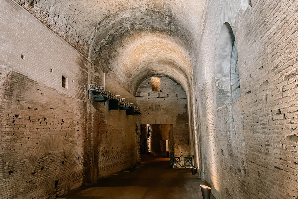 An ancient brick corridor with arched ceilings, illuminated by warm lights, featuring a distant gated archway.